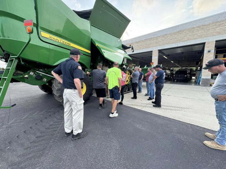 A group of people stand around a large green John Deere combine harvester, parked in front of a workshop. Some are inspecting the machinery, while others are engaged in conversation. The setting appears to be an outdoor agricultural equipment demonstration or workshop.