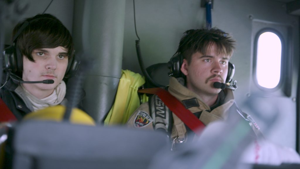 Two firefighters wearing headsets and safety gear sit inside a vehicle, possibly a fire truck or helicopter, looking focused and alert. One wears a red harness, both appear attentive to their tasks.