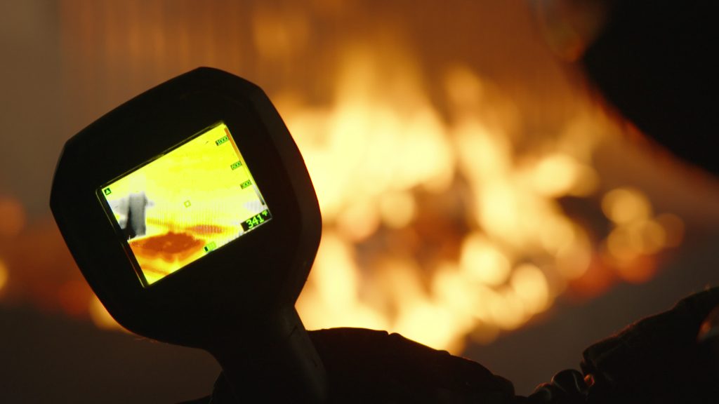 A person holds a thermal imaging camera displaying a heat map while facing bright, intense flames in the background.