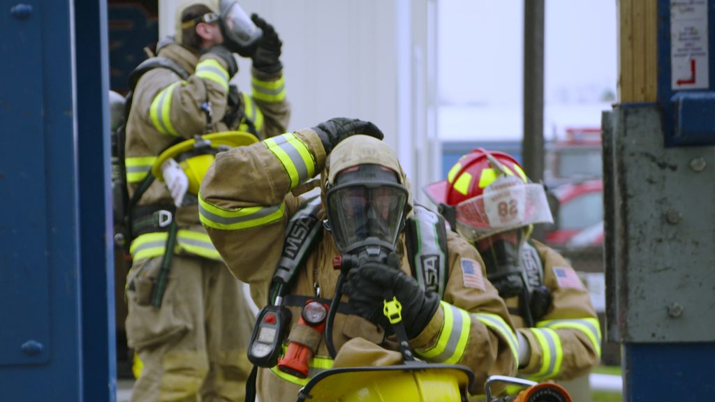 Three firefighters in full protective gear and helmets prepare for action, adjusting their masks and equipment. One crouches in the foreground while two others stand behind, all appearing focused and ready.