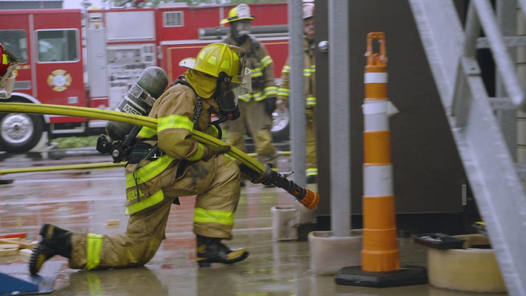 A firefighter in yellow gear kneels on wet pavement, holding a hose near a building. Other firefighters and a red fire truck are visible in the background, with orange traffic cones nearby.