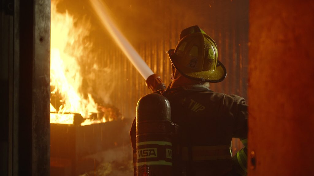 A firefighter in full gear sprays water from a hose onto intense flames inside a burning structure at night.