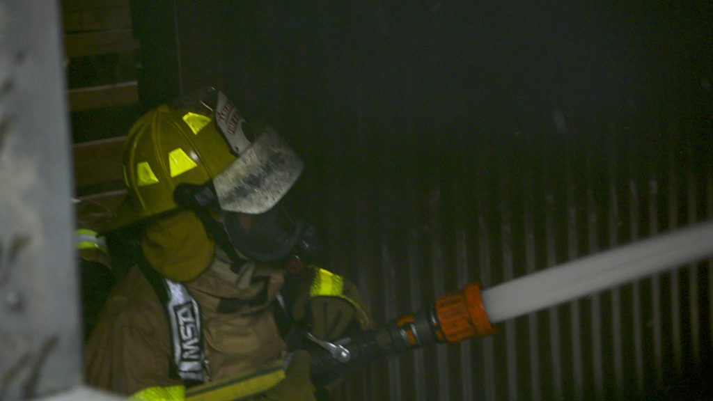 A firefighter in full protective gear and helmet sprays water from a fire hose in a smoky, dark environment, likely battling a fire indoors.