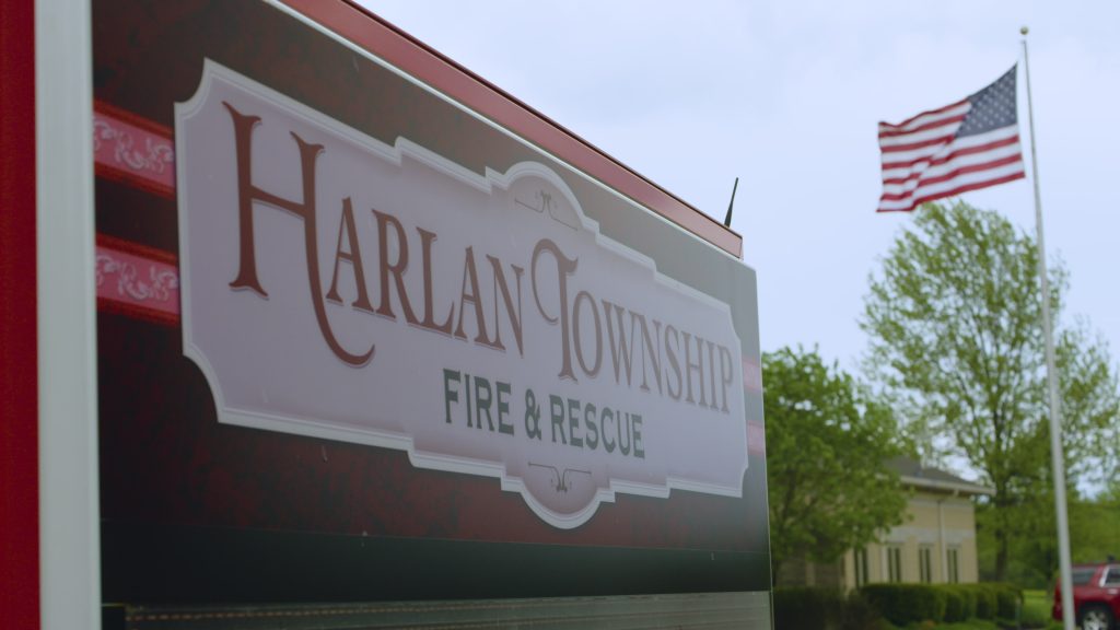 A sign for Harlan Township Fire & Rescue is in the foreground, with an American flag and some trees and a building visible in the background under a cloudy sky.