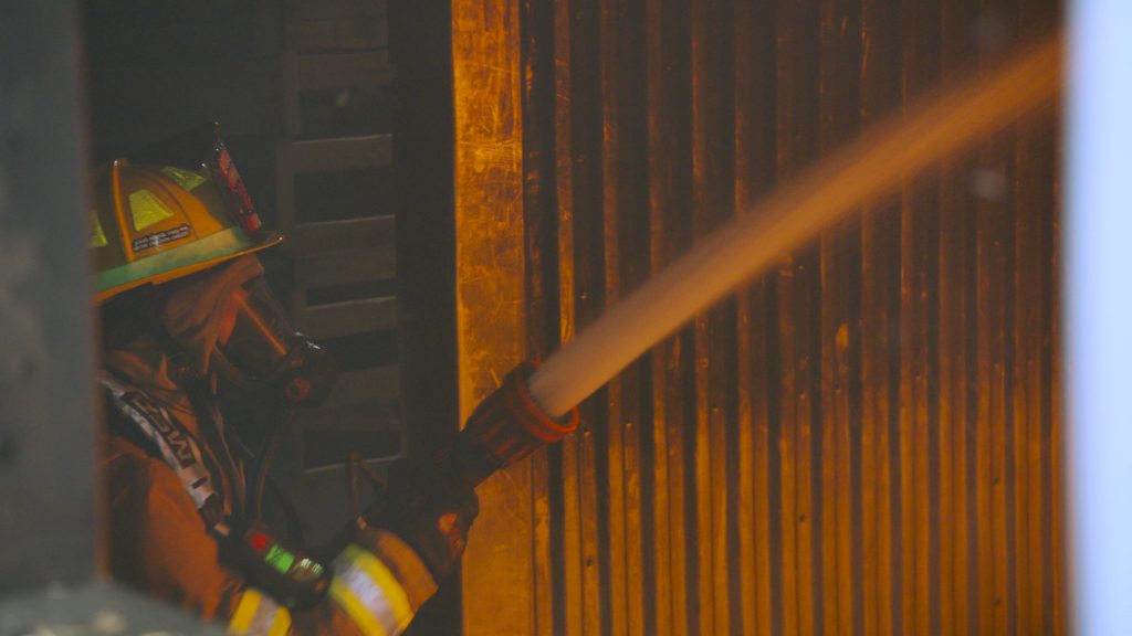 A firefighter in full gear sprays water from a hose onto a burning metal wall, with orange flames reflecting off the surface.