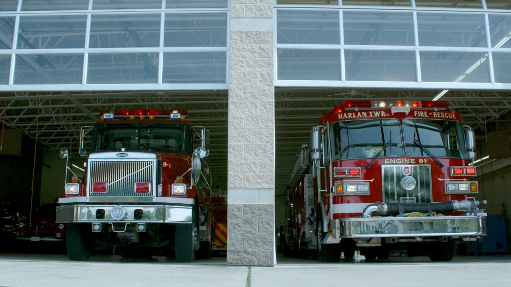 Two red fire trucks are parked side by side inside a fire station garage with the doors partially open, revealing the front of the vehicles. The words "Fire-Rescue" and "Engine 81" are visible on one truck.